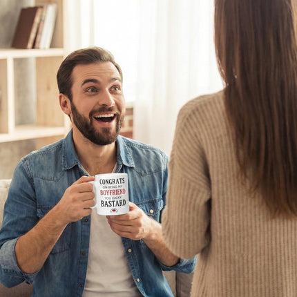 A happy man holding a coffee mug with a surprised expression, looking at a woman standing in the foreground. He is receiving a funny novelty gift mug