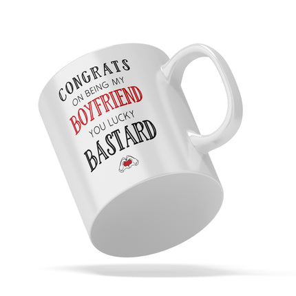 Angled studio shot of a white novelty mug floating against a white background. The text on the mug reads CONGRATS ON BEING MY BOYFRIEND YOU LUCKY BASTARD