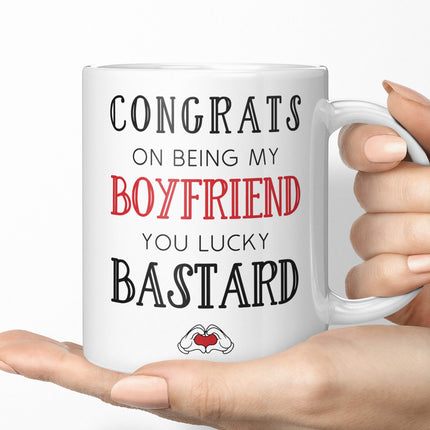 Close-up of a person's hands holding a white coffee mug. The mug displays the humorous romantic gift message CONGRATS ON BEING MY BOYFRIEND YOU LUCKY BASTARD