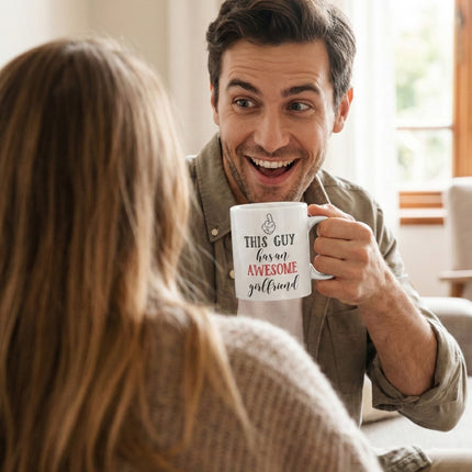 A lifestyle shot of a smiling man holding a coffee mug that reads "This Guy Has An Awesome Girlfriend," looking excitedly at his partner in a cozy home setting.