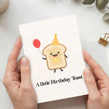 Greeting card with a cartoon toast character and 'A little Birthday Toast' text, held by hands on a light background.