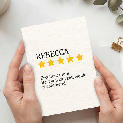 A top-down view of a pair of hands holding a personalized Mother's Day greeting card with a textured, off-white finish. The card features the name "REBECCA" in black serif text above five yellow stars.