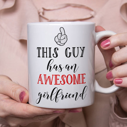 A close-up view of hands holding a white ceramic coffee mug featuring the phrase "This Guy Has An Awesome Girlfriend" in mixed black and red typography with a cartoon pointing finger graphic.
